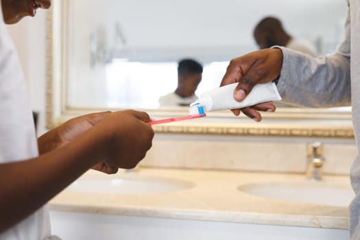 African american father and son putting toothpaste on toothbrush in bathroom. family spending time at home.