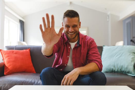 Happy caucasian man sitting on couch having video call in living room, smiling and waving. keeping in touch, leisure time at home with communication technology.