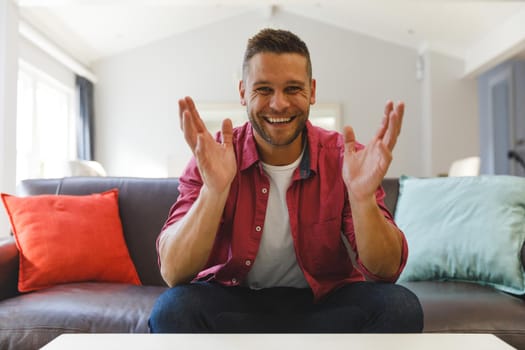 Happy caucasian man sitting on couch having video call in living room, smiling and gesturing. keeping in touch, leisure time at home with communication technology.