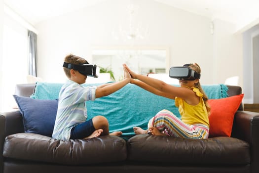 Caucasian brother and sister sitting on couch and using vr headsets in living room. childhood leisure time, fun and discovery at at home using technology.