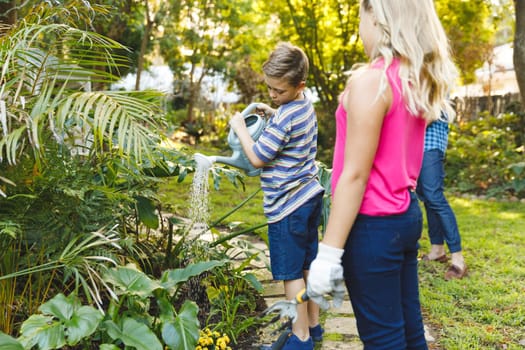 Caucasian son and daughter watering plants in garden with mother and father in background. family enjoying leisure time together gardening at home.