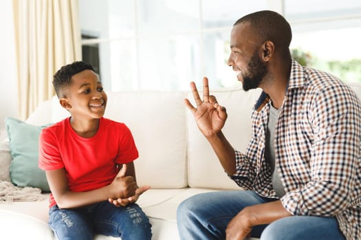 Happy african american father with son sitting on couch in living room talking sign language. father and son communicating without words.