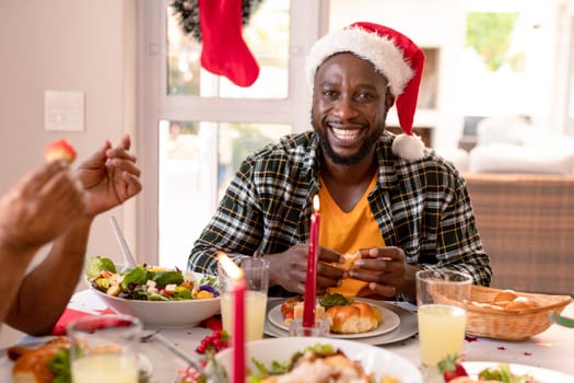 Happy african american man wearing santa hat sitting at christmas table. family christmas time and festivity together at home.