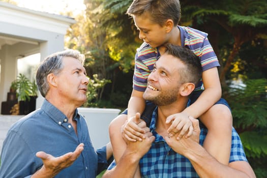 Portrait of caucasian grandfather and smiling adult son with grandson on his shoulders in garden. multi generation family enjoying leisure time together at home.