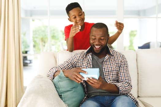 African american father opening gift from his son and smiling in living room. family spending time at home, father son relationship.