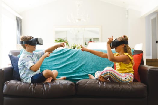 Caucasian brother and sister sitting on couch and using vr headsets in living room. childhood leisure time, fun and discovery at at home using technology.