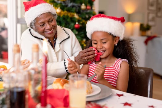 Happy african american grandmother cleaning granddaughter face at christmas table. family christmas time and festivity together at home.