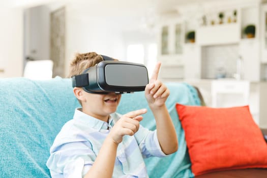 Caucasian boy sitting on couch and using vr headset in living room. childhood leisure time, fun and discovery at at home using technology.