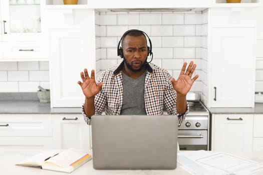 Gesturing african american man sitting in kitchen making video call using laptop and headset. remote working from home with technology.