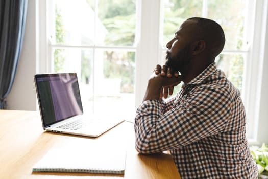 African american man sitting at table in dining room, working remotely using laptop. flexible working from home with technology.