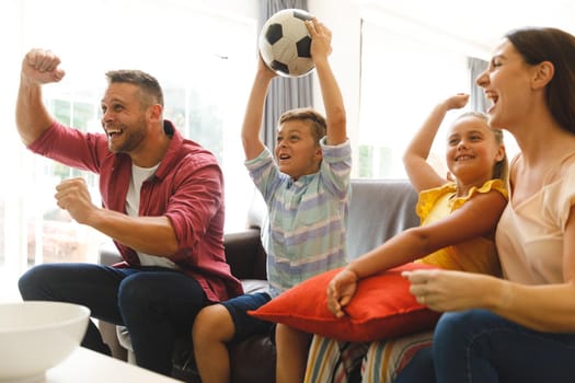 Excited caucasian parents on couch with daughter and son watching football match on tv and cheering. family entertainment and leisure time together at home.