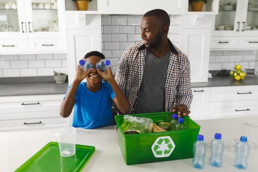 Happy african american son sorting recycling with father in kitchen, playing with plastic bottles. family spending time at home.