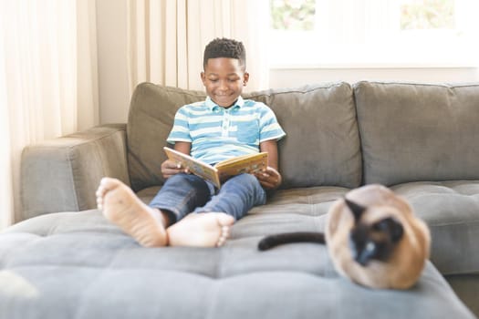 Smiling african american boy reading book and sitting on couch with cat in living room. spending time alone at home.