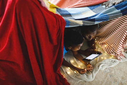 Happy african american father and son lying in blanket fort, using tablet. family spending time at home.