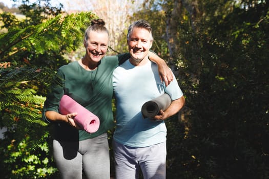 Portrait of happy senior caucasian couple holding yoga mats, looking to camera in sunny garden. healthy retirement lifestyle, spending time at home.