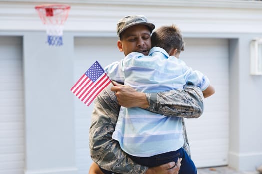 Smiling caucasian male soldier with son outside house holding american flags. soldier returning home to family.