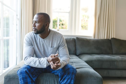 Thoughtful african american man sitting on couch looking out of window in living room. spending time alone at home.