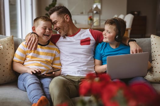 Portrait of caucasian father and two sons with laptop and digital tablet smiling at home. fatherhood and love concept