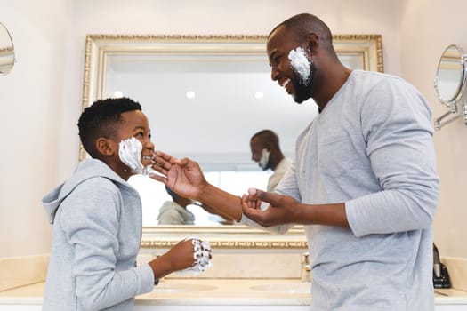 African american father with son having fun with shaving foam in bathroom. family spending time at home.