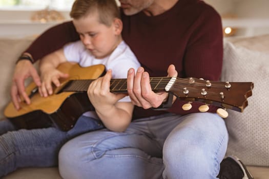 Mid section of caucasian father teaching his son to play guitar sitting on the couch at home. childhood and hobby concept