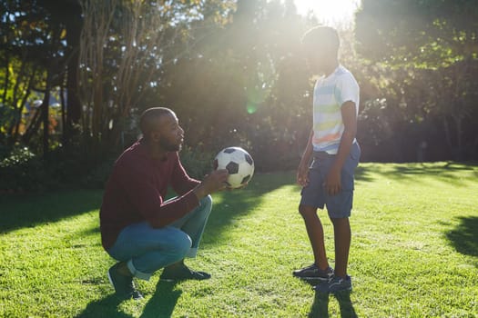 African american father with son having fun and playing with football and talking in sunny garden. family spending time at home.