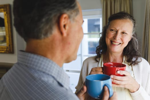 Happy senior caucasian couple standing next to window, holding cups. retirement lifestyle, spending time at home.