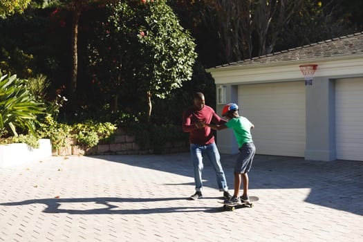 African american father smiling and helping son balancing on skateboard in garden. family spending time at home.