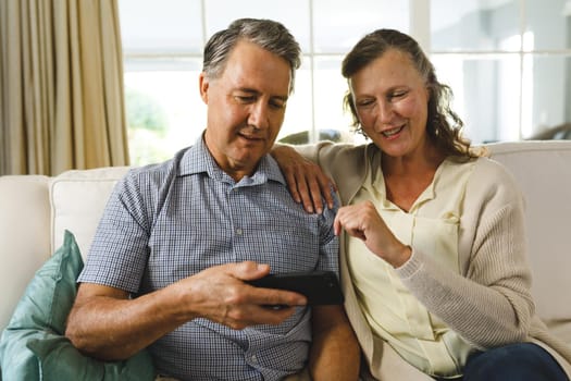Happy senior caucasian couple in living room, sitting on sofa, using smartphone. retirement lifestyle, spending time at home.