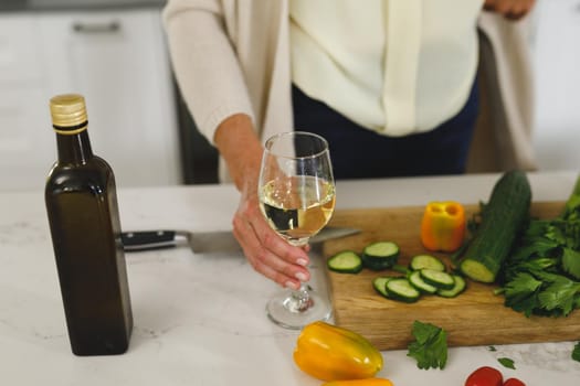 Midsection of senior caucasian woman in modern kitchen, holding glass of wine. retirement lifestyle, spending time at home.