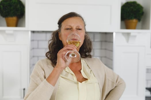 Senior caucasian woman in modern kitchen, holding glass of wine and drinking. retirement lifestyle, spending time at home.