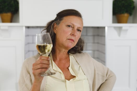 Portrait of senior caucasian woman in modern kitchen, holding glass of wine, looking to camera. retirement lifestyle, spending time at home.