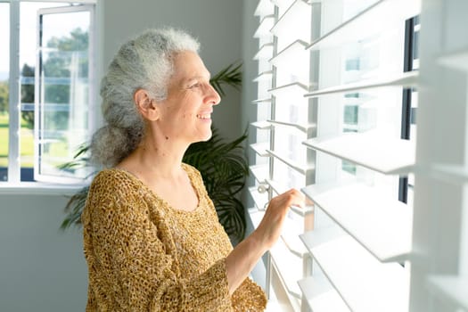 Smiling caucasian senior woman standing at window, looking into distance. healthy retirement lifestyle at home.