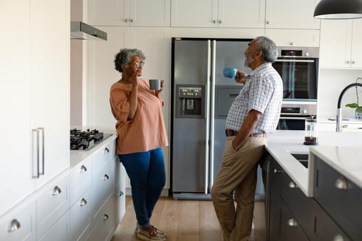 Happy african american senior couple standing in kitchen, holding mugs with coffee and talking. healthy retirement lifestyle at home.