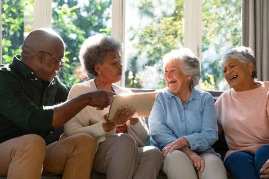 Three happy diverse senior woman and african american male friend sitting on sofa and using tablet. retirement lifestyle relaxing at home with technology.