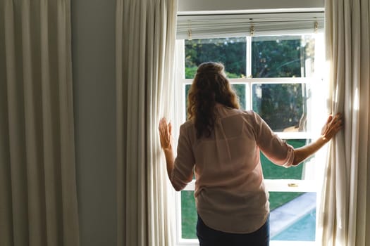 Thoughtful senior caucasian woman in bedroom, standing next to window, opening curtains. retirement lifestyle, spending time alone at home.