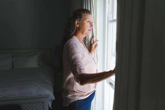 Thoughtful senior caucasian woman in bedroom, standing next to window, widening curtains. retirement lifestyle, spending time alone at home.