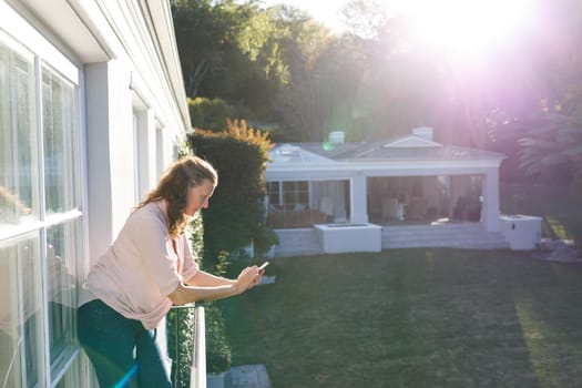 Happy senior caucasian woman using smartphone, standing on sunny balcony. retirement lifestyle, spending time alone at home.