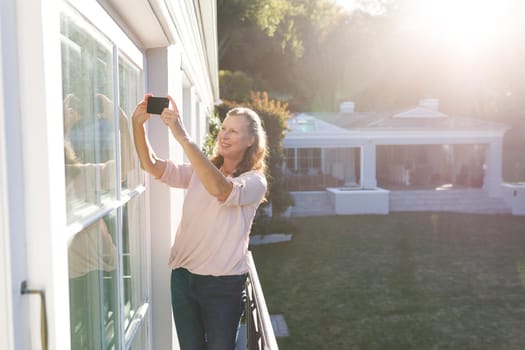 Happy senior caucasian woman taking selfies, standing on sunny balcony. retirement lifestyle, spending time alone at home.