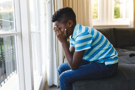 Sad african american boy covering his face and sitting at window in living room. spending time alone at home.