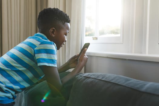 Happy african american boy kneeling on couch using smartphone in living room. spending time alone at home with technology.