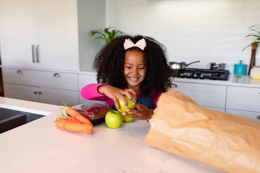 Happy african american girl unpacking groceries in kitchen. childhood, leisure and spending time at home.