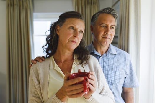 Happy senior caucasian couple standing next to window, holding cups. retirement lifestyle, spending time at home.