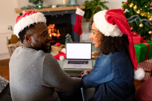 Happy african american couple wearing santa hats, using laptop with copy space on screen. christmas, festivity and communication technology.