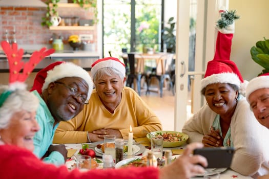 Diverse group of happy senior friends in holiday hats celebrating christmas together, taking selfie. christmas festivities, celebrating at home with friends.