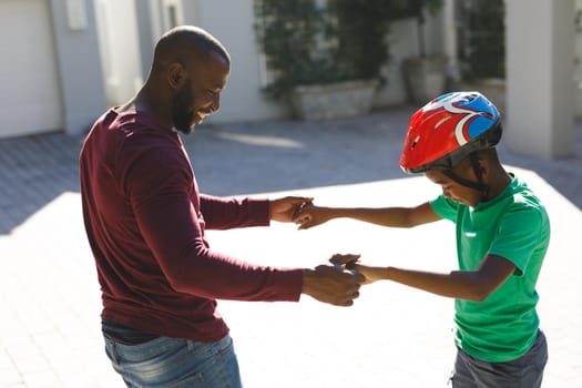 African american father smiling and helping son balancing on skateboard in garden. family spending time at home.