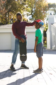 African american father with son smiling and preparing before skateboarding in garden. family spending time at home.