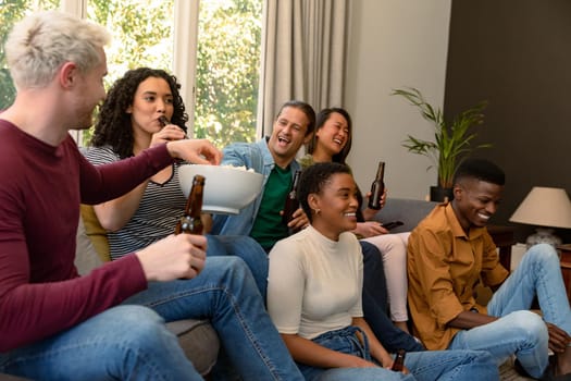 Group of happy diverse female and male friends watching tv and drinking beer together at home. socialising with friends at home.