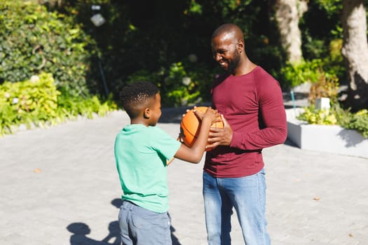 African american father with son smiling and playing basketball in garden. family spending time at home.