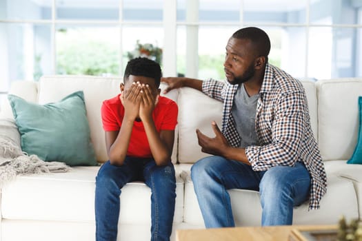 Sad african american son sitting on couch covering face listening to father talking in living room. family spending time at home, father son relationship.