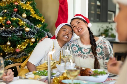 Happy diverse female friends in santa hats embracing, celebrating christmas with friends at home. christmas festivities, celebrating at home with friends.